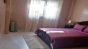 Quarto com cama, mesa de cabeceira, luminária, radiador e janela com cortinas.