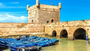 Um edifício histórico de pedra com uma torre e arcos, rodeado por barcos azuis em águas calmas.