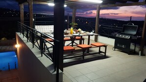 An outdoor dining area with a pool, a grill, and a view of the city at dusk.