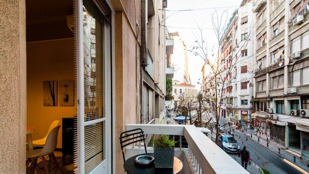 A balcony with a table and chairs, overlooking a street with buildings and shops. - We saw athens from 1a (Αθήνα)