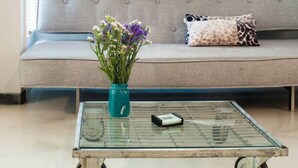 A room with a sofa, a glass coffee table, and a window with blinds. - We saw athens from 1a (Αθήνα)