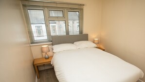A neatly made bed with white bedding, two bedside lamps, and a window with blinds. - Celtic Bunkhouse Hostel (Merthyr Tydfil)