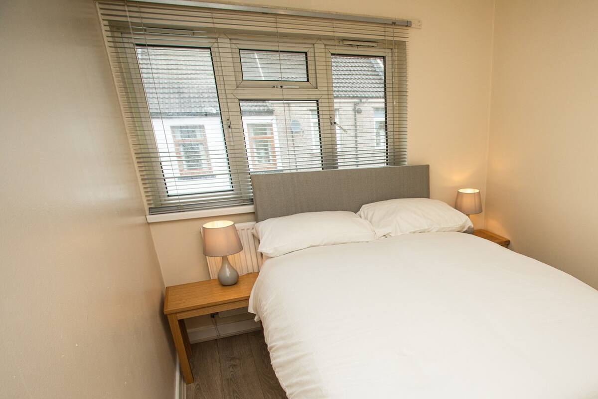 A neatly made bed with white bedding, two bedside lamps, and a window with blinds.