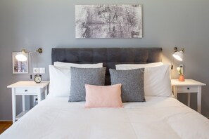 A neatly made bed with a grey headboard, white bedding, and multiple pillows. There are bedside tables with lamps and a clock on one side.