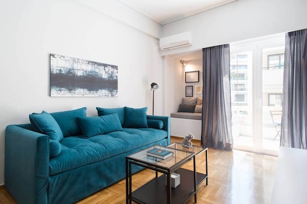 A modern living room with a teal sofa, a glass coffee table, and a large window with curtains.