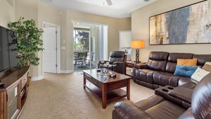 A spacious living room with a leather sofa, a coffee table, and a TV. There is a ceiling fan, a painting on the wall, and a plant. - LUXE DISNEY HOME, POOL W/ SPA, GAME ROOM,  FREE NETFLIX & WIFI  (Davenport)