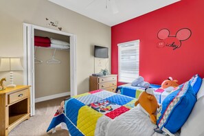 A hotel room with a red accent wall, a bed with a colorful quilt, a Mickey Mouse decal, a wardrobe, and a bedside table with a lamp.