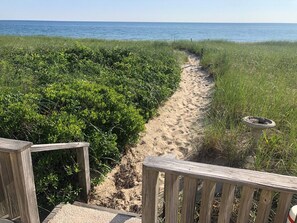 A sandy path leading to the beach, surrounded by greenery, with a wooden deck and a bird bath. - 233 Phillips Rd (Sandwich)