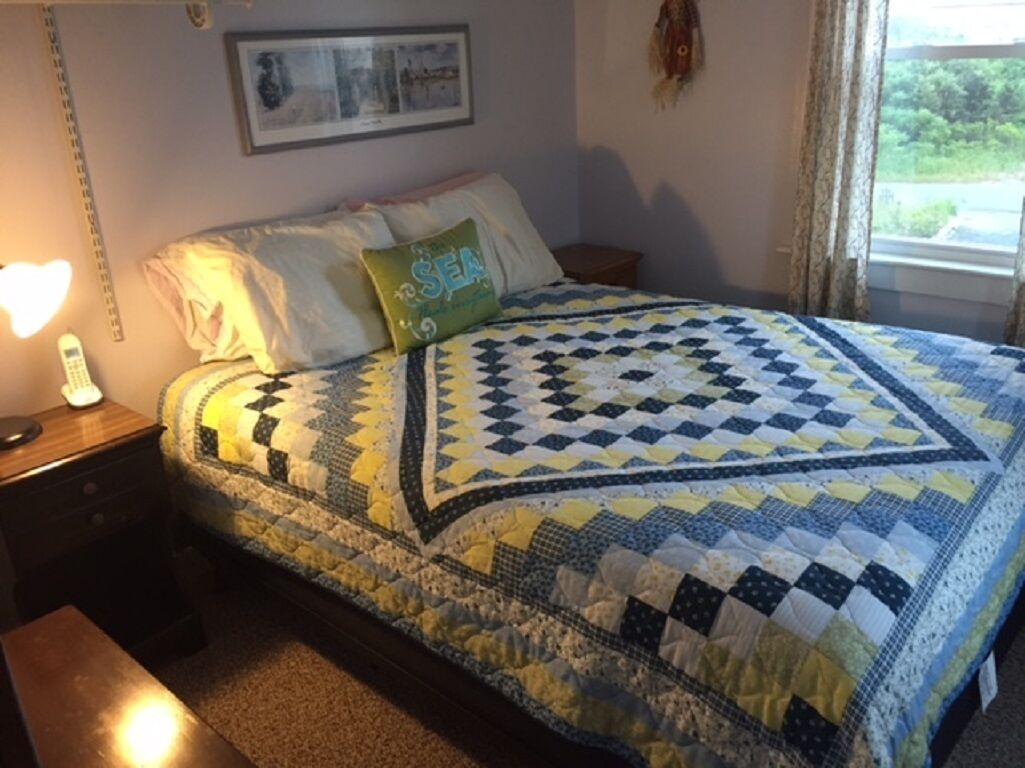 A bedroom with a quilted bedspread, a bedside lamp, a framed picture on the wall, and a window with curtains.