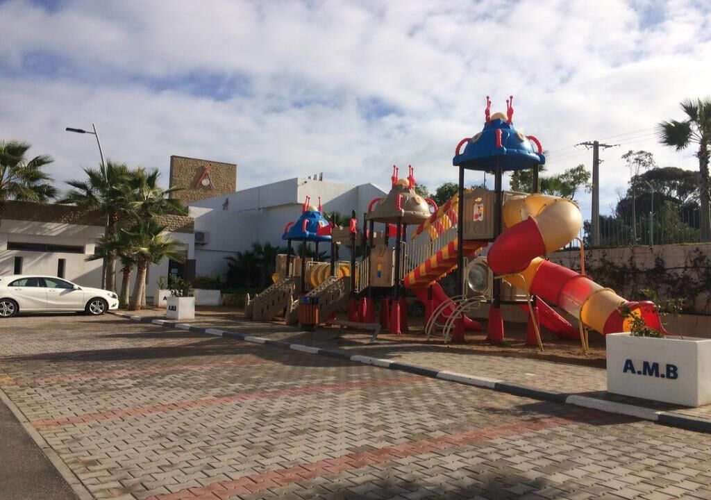 A playground with slides and climbing structures, a white car parked nearby, and a building with a sign reading "A.M.B" in the foreground.