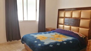 A bedroom with a bed, a nightstand, a window with curtains, and a headboard with a decorative panel. - Nice apartment in Essaouira 4267 (Essaouira)