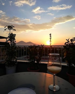 A table with a glass of drink and potted plants on a balcony with a sunset view.