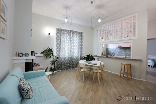 A modern living room with a light blue sofa, a dining area with a round table, and a kitchen with a window.