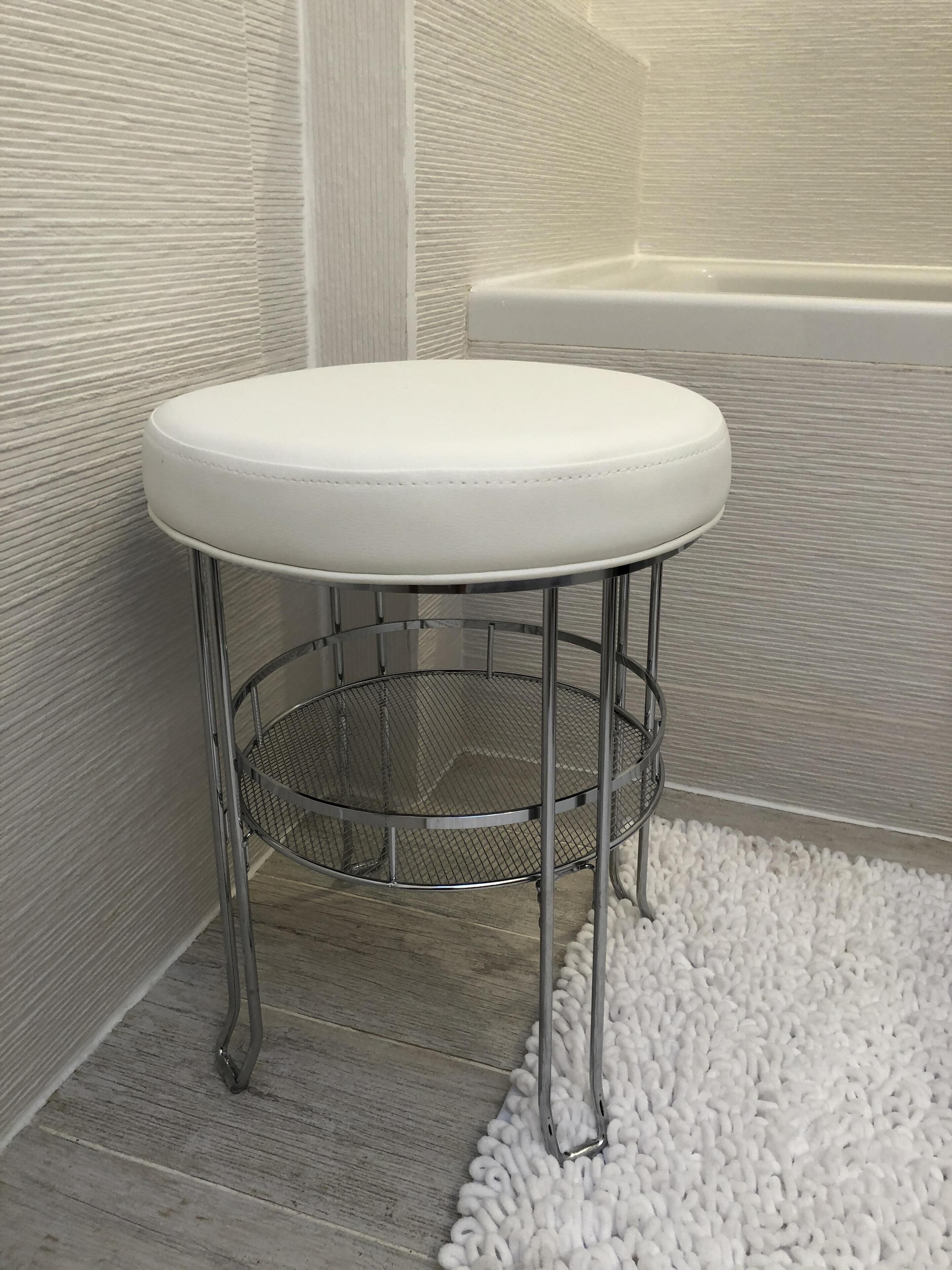 Un taburete blanco acolchado con una base de mimbre metálico, colocado sobre una alfombra blanca con textura en un baño con una bañera al fondo.