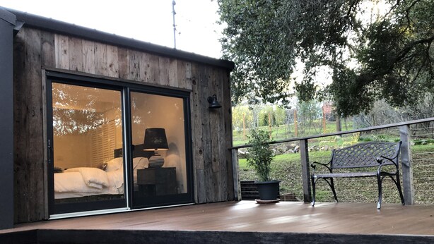 A modern building with a large sliding glass door leading to a bedroom with a bed and a lamp. There is a wooden deck with steps and a bench.
