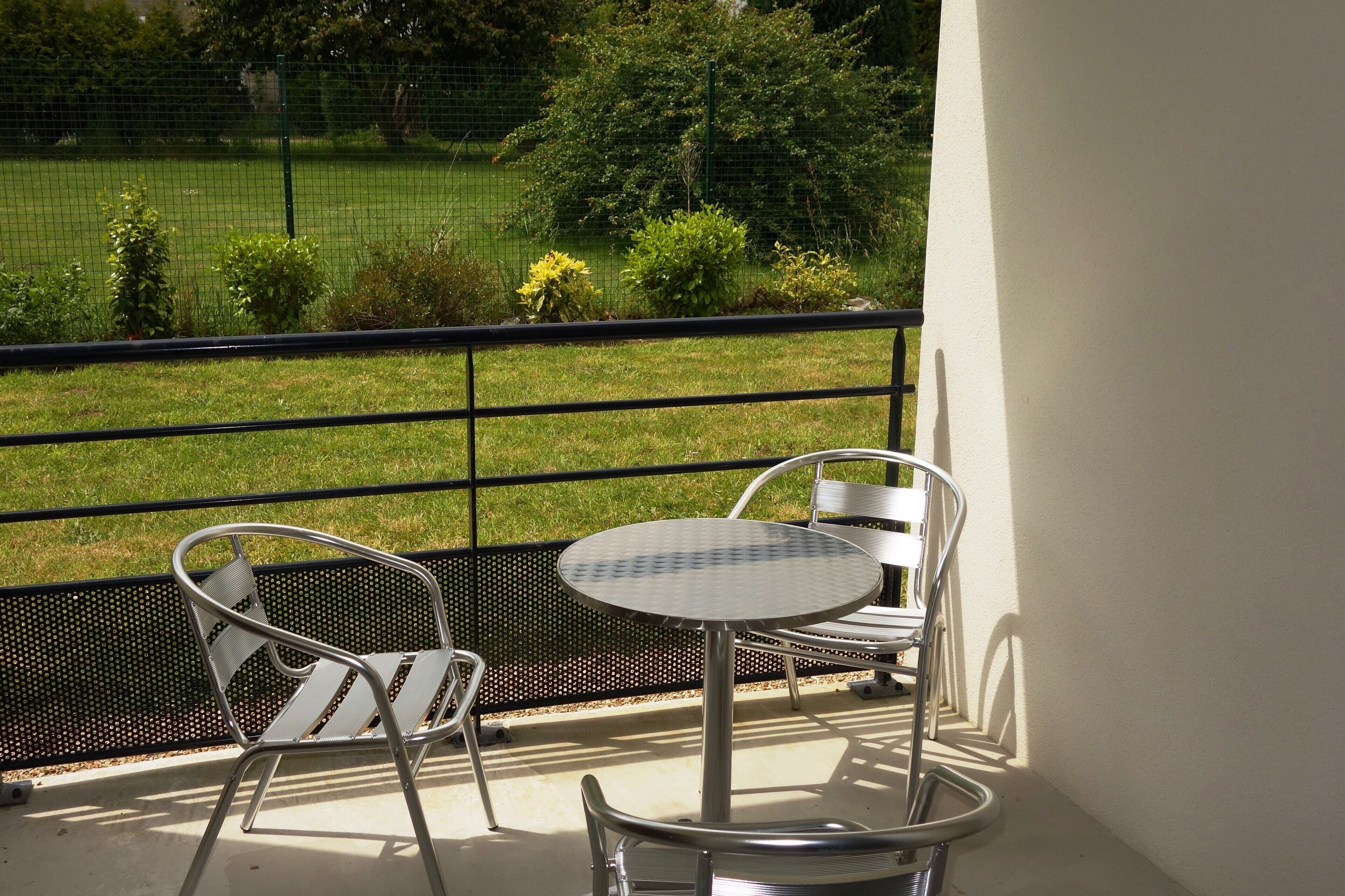 A balcony with a metal table and two chairs, overlooking a grassy area with a fence and trees.