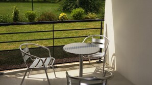 Ein Balkon mit einem Metalltisch und zwei Stühlen, mit Blick auf eine Rasenfläche mit Zaun und Bäumen.