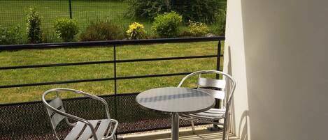 A balcony with a metal table and two chairs, overlooking a grassy area with a fence and trees.