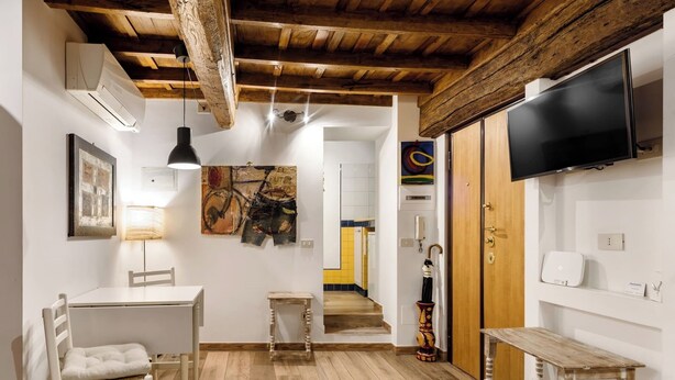 A room with a wooden ceiling, white walls, a TV mounted on the wall, a table with a chair, and a staircase leading to another room.