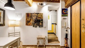 A room with a wooden ceiling, a white table with a chair, a small wooden stool, a lamp, and a door with a decorative umbrella.