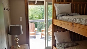 Une chambre avec un lit superposé, une table avec une lampe et une vue sur une terrasse en bois et la verdure environnante.