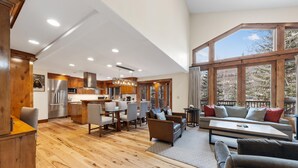 A spacious living area with a wooden floor, a dining table with chairs, a sofa, and a large window offering a view of trees.