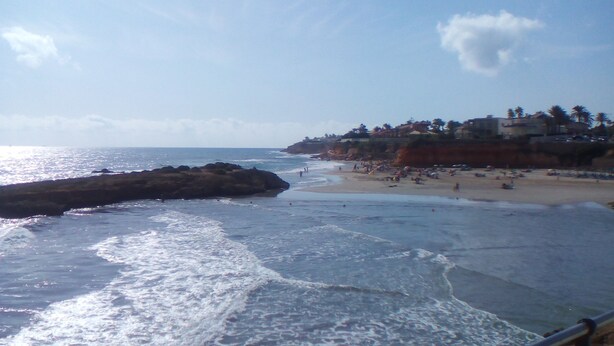 Una playa con olas, un promontorio rocoso y un edificio con palmeras.
