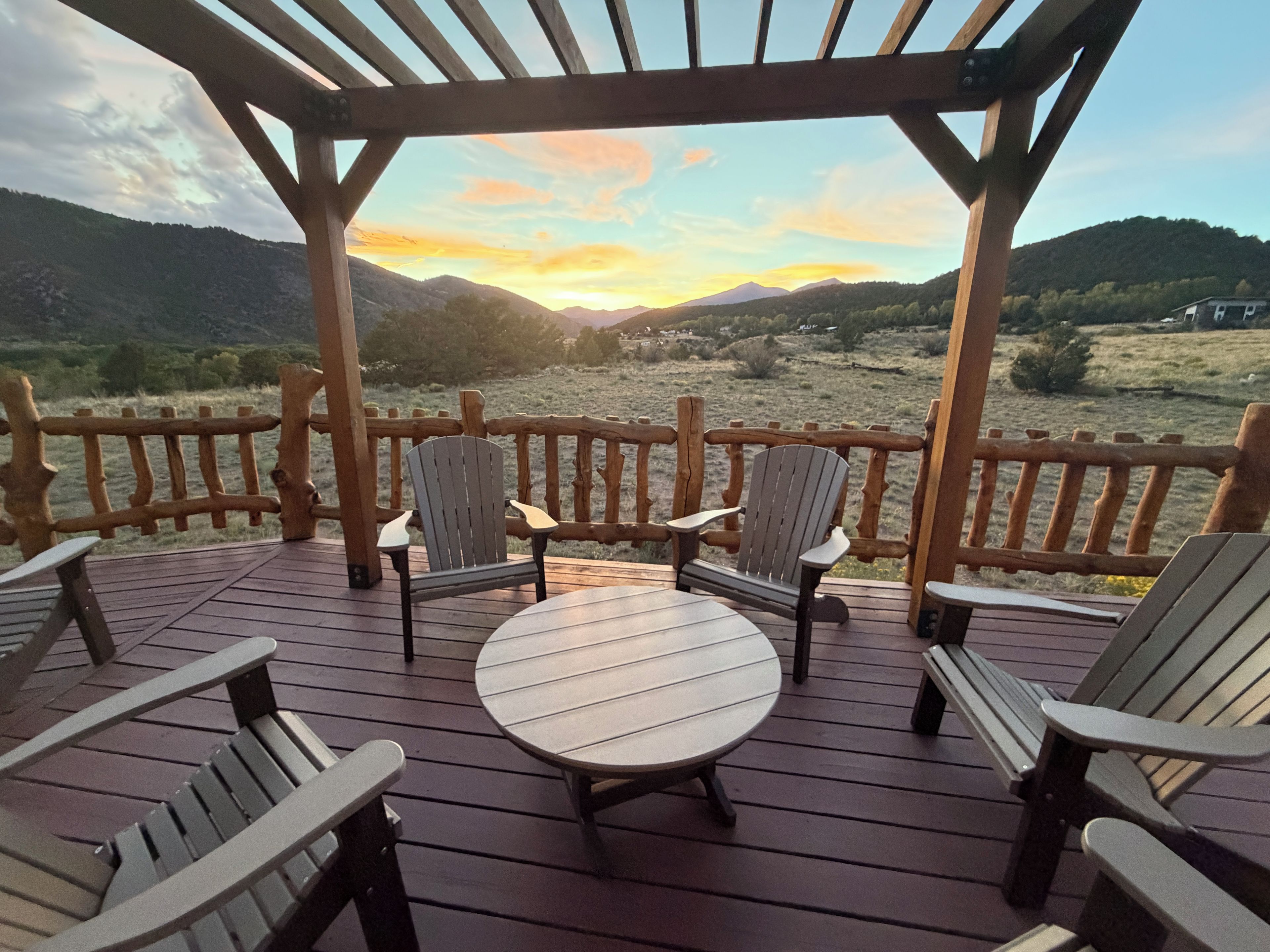 Een houten terras met Adirondack-stoelen en een ronde tafel, met uitzicht op een bergachtig landschap.