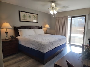A hotel room with a large bed, two bedside lamps, a ceiling fan, a window with curtains, and a framed picture on the wall.