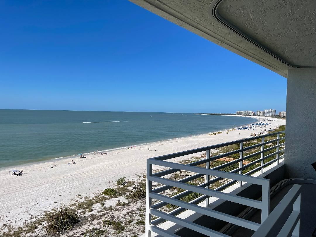 A beach view from a balcony with a railing, overlooking a sandy beach and the ocean.