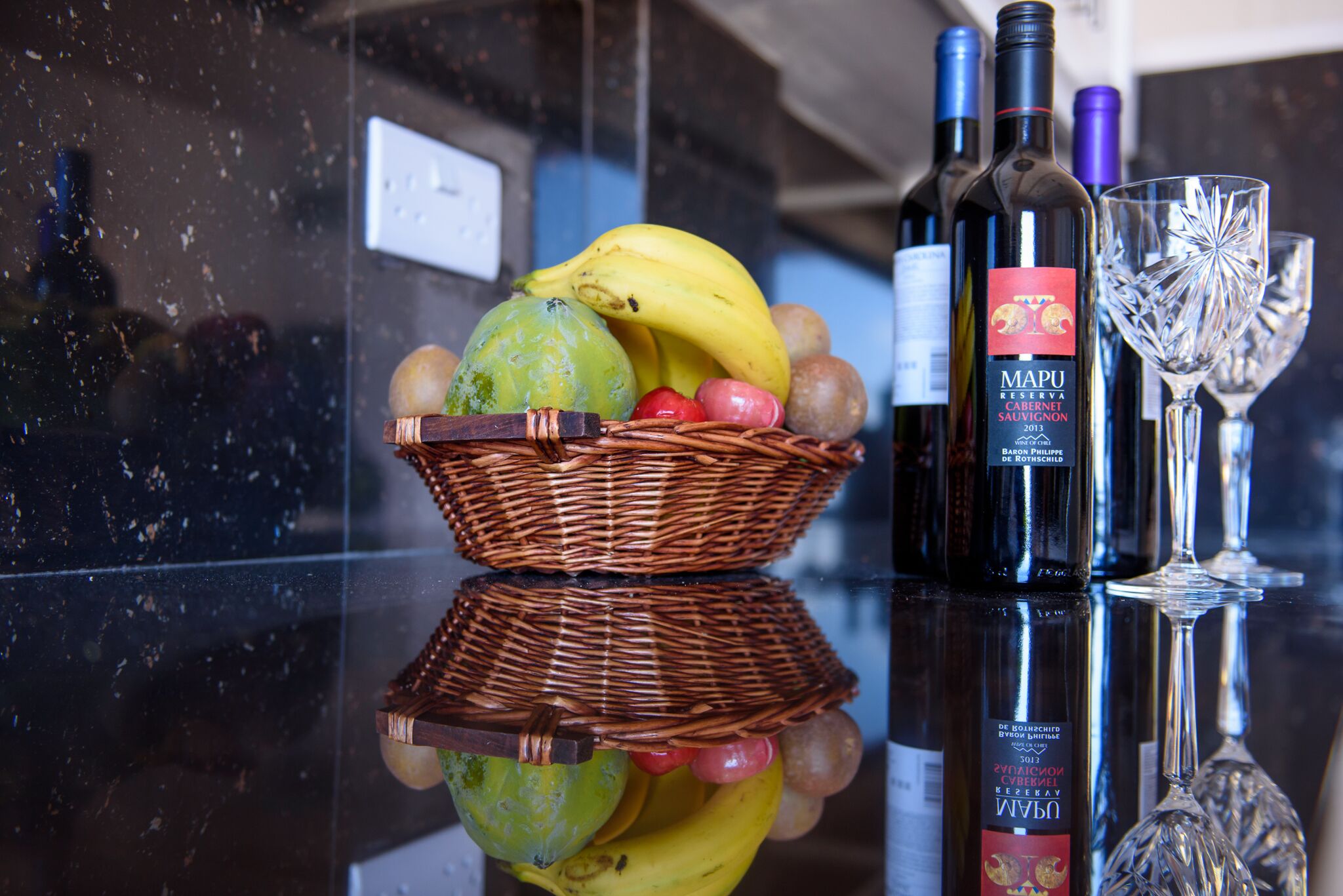 A reflective surface with a wicker basket containing fruits, a bottle of wine, and a wine glass.
