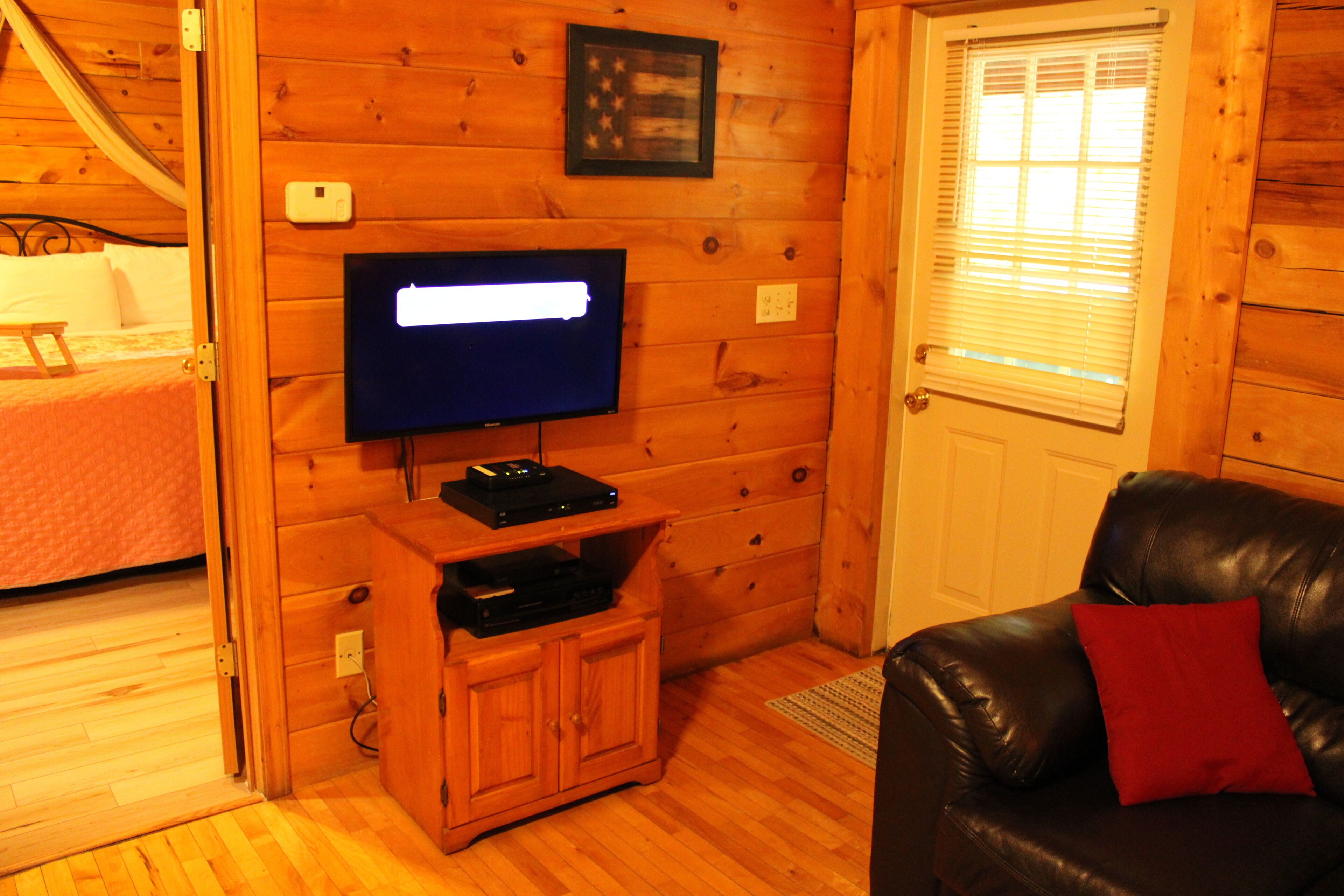 Interior d'una cabana de fusta amb una televisió sobre un suport de fusta, una cadira de cuir amb coixí vermell i un llit amb roba de llit blanca.