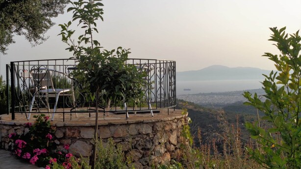 Uma plataforma de pedra com guarda-corrimão de metal, uma cadeira e uma mesa com vista panorâmica para uma cidade e montanhas.