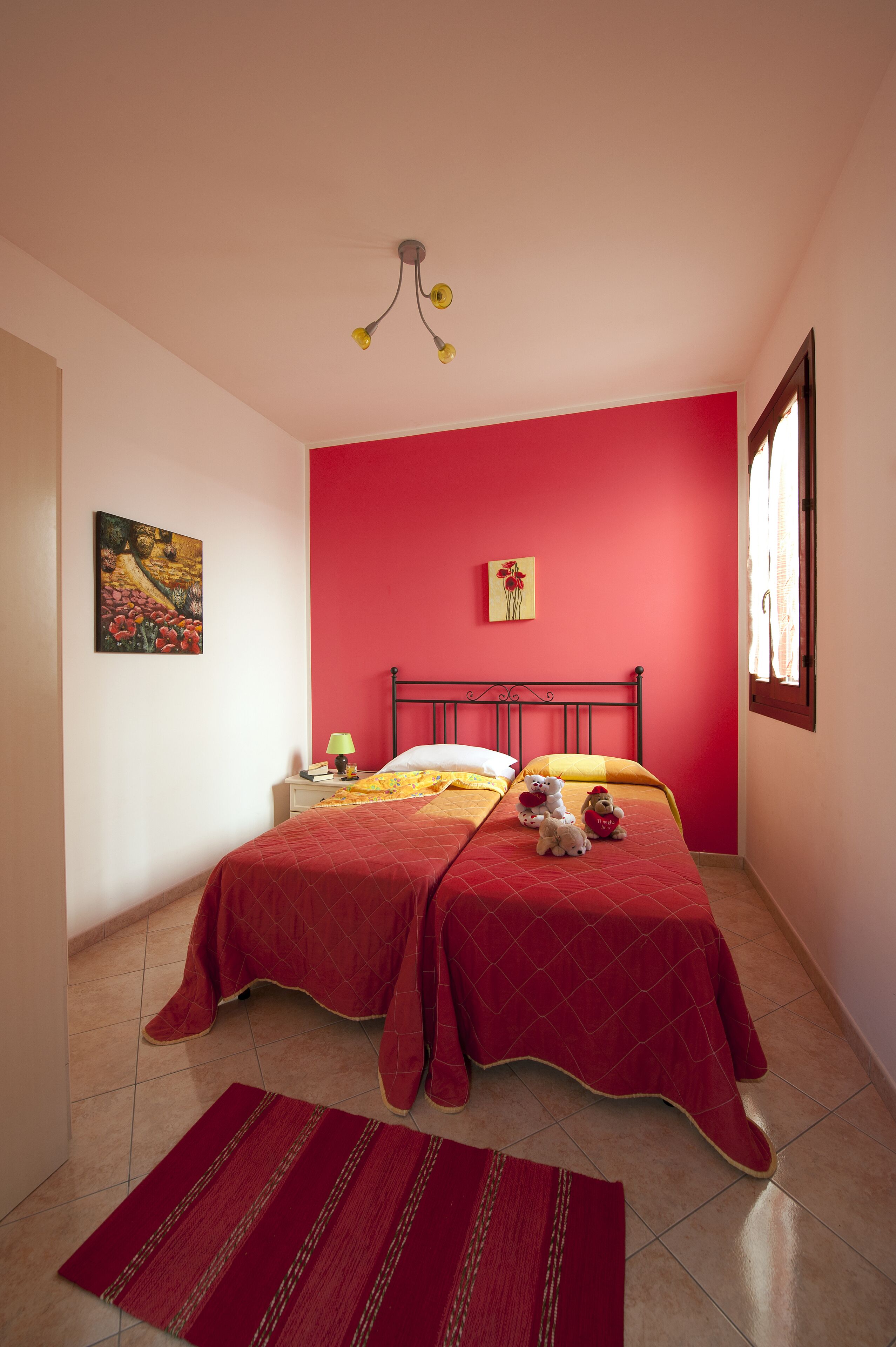 Een slaapkamer met twee bedden, een rode muur, een schilderij en een raam met gordijnen.