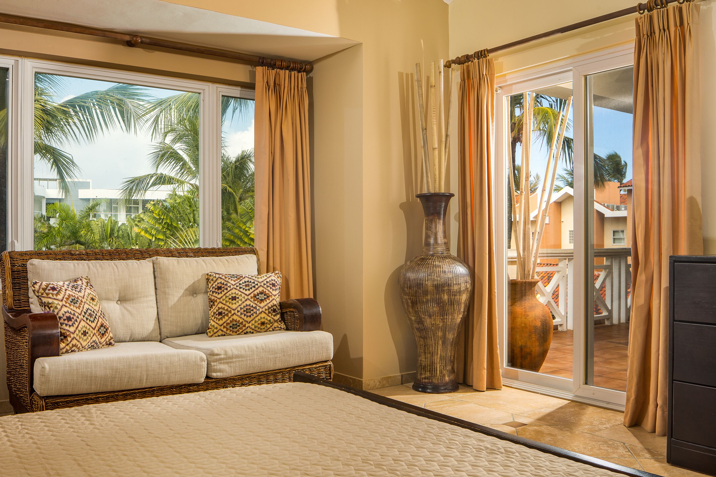 A bedroom with a large bed, a wicker sofa with patterned cushions, a large vase, and a sliding glass door leading to a balcony with palm trees.