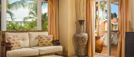 A bedroom with a large bed, a wicker sofa with patterned cushions, a large vase, and a sliding glass door leading to a balcony with palm trees.