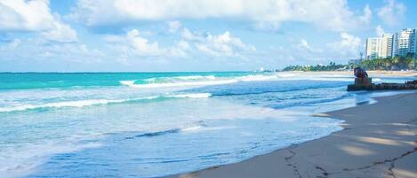Una playa con olas, una escultura y edificios al fondo.