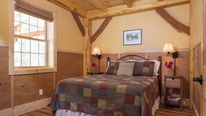 A bedroom with a wooden ceiling, a large window, a bed with a quilt, two bedside lamps, and a small nightstand with books. - Private pet-friendly cabin in the woods w/ private campfire pit & screened porch (Ripton)