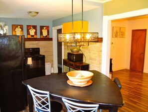 A kitchen with a black refrigerator, a microwave, and a stove. There is a dining table with a bowl on it and two white chairs. A hanging light fixture is above the table.
