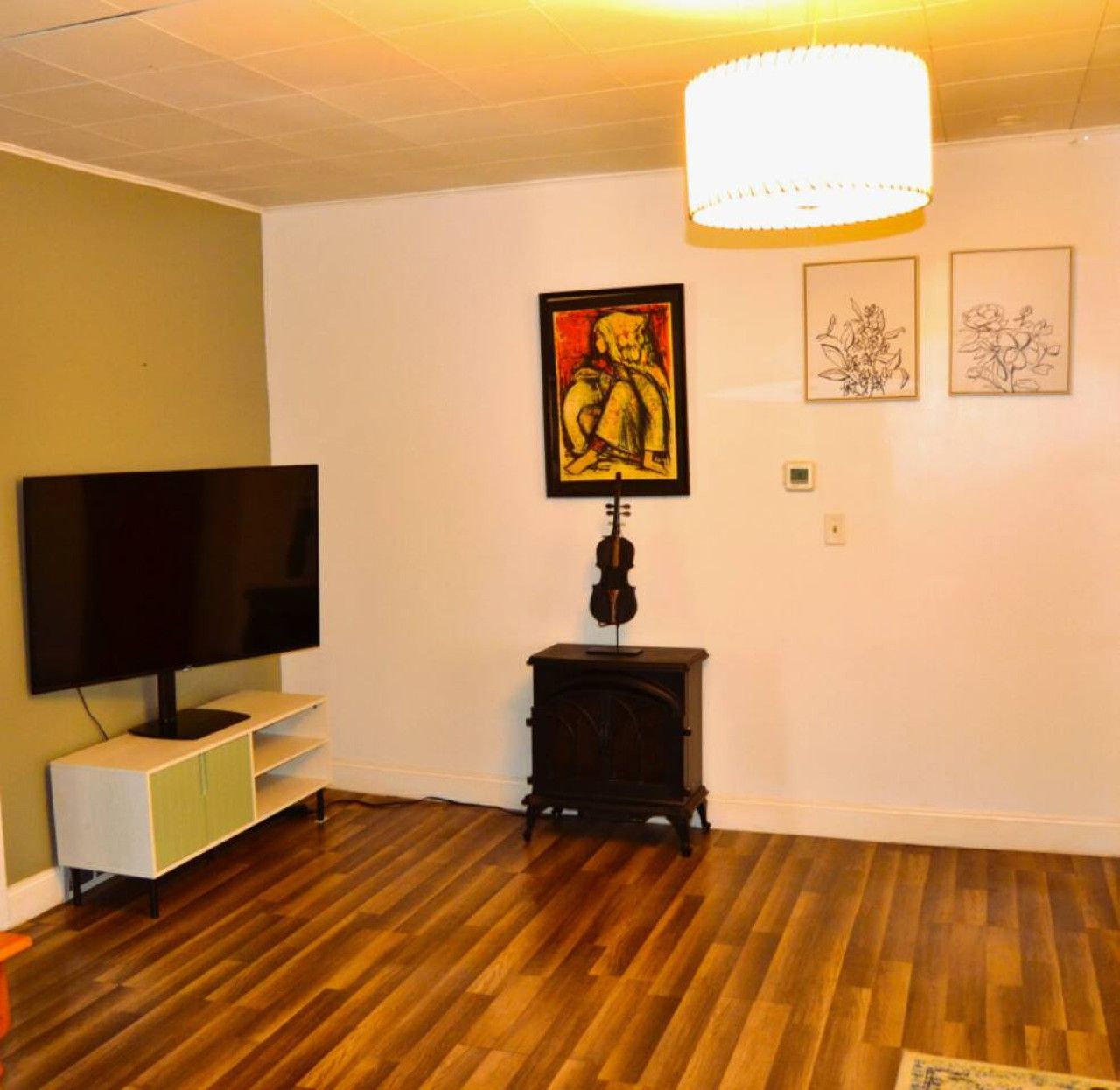 A room with a flat-screen TV on a white stand, a wooden floor, a black music stand with a violin, and three framed artworks on the wall.