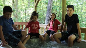 Four individuals sitting in a hot tub surrounded by a wooden deck and trees. - Spacious Cabin sleeps 8! (Hico)
