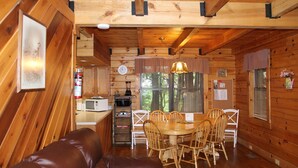 A cozy cabin interior with a wooden dining table, chairs, a brown leather sofa, a kitchen area with appliances, and a large window with blinds. - Spacious Cabin sleeps 8! (Hico)