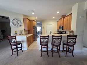 A kitchen with wooden cabinets, a stainless steel refrigerator, a clock on the wall, and three bar stools.
