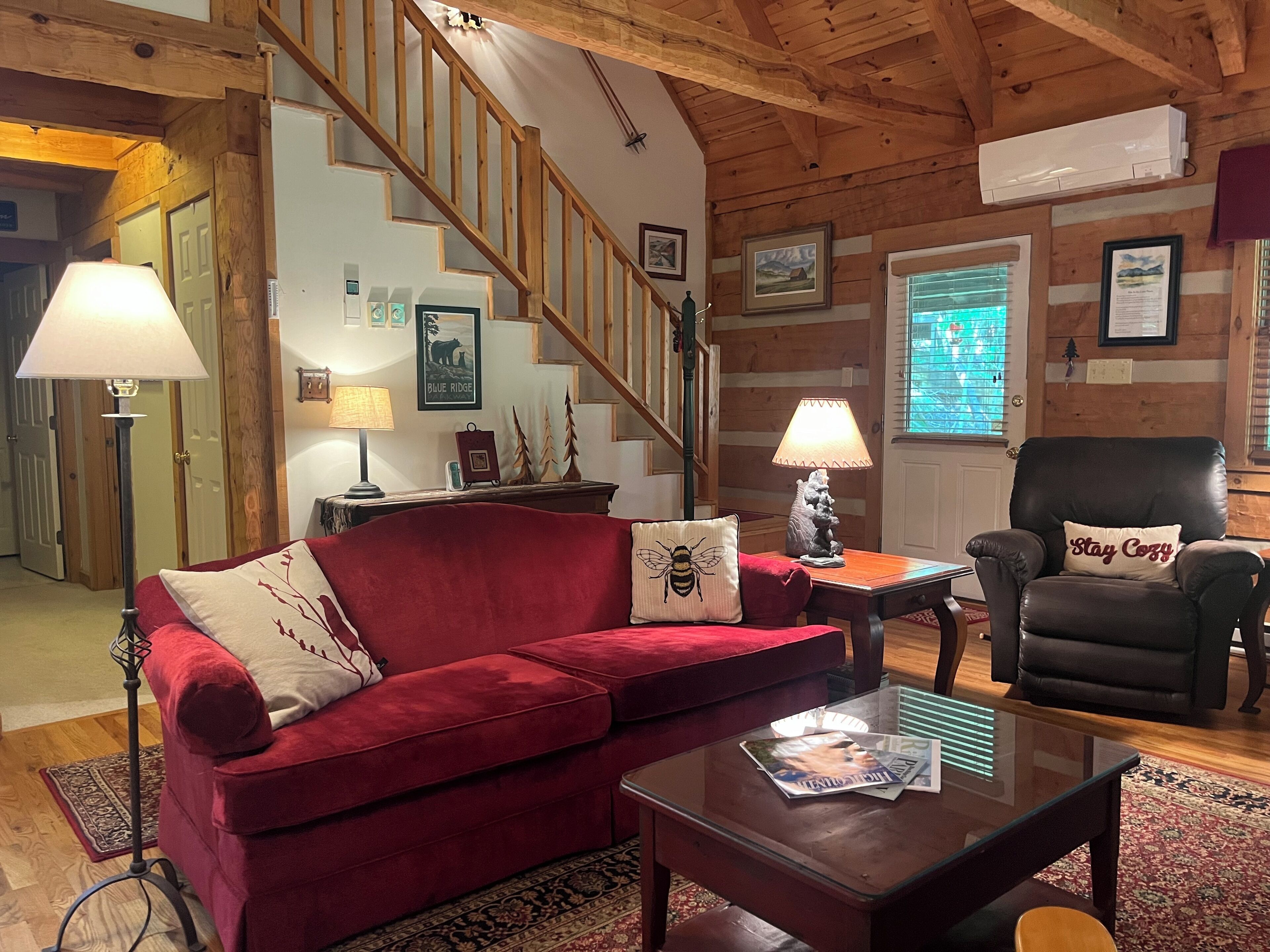 A cozy living room with a red sofa, a glass coffee table, a black leather armchair, and a wooden staircase.