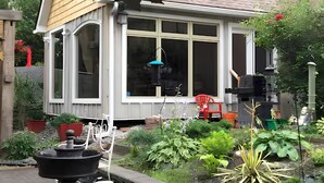 Une petite maison avec une grande fenêtre, un jardin où poussent différentes plantes et une chaise rouge à l’extérieur.