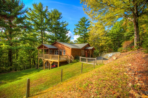 A cabin with a deck and a fenced area, surrounded by trees and a grassy slope. - DOWNTOWN BLOWING ROCK(1.5mi) Three Bears Hot tub (Lenoir)