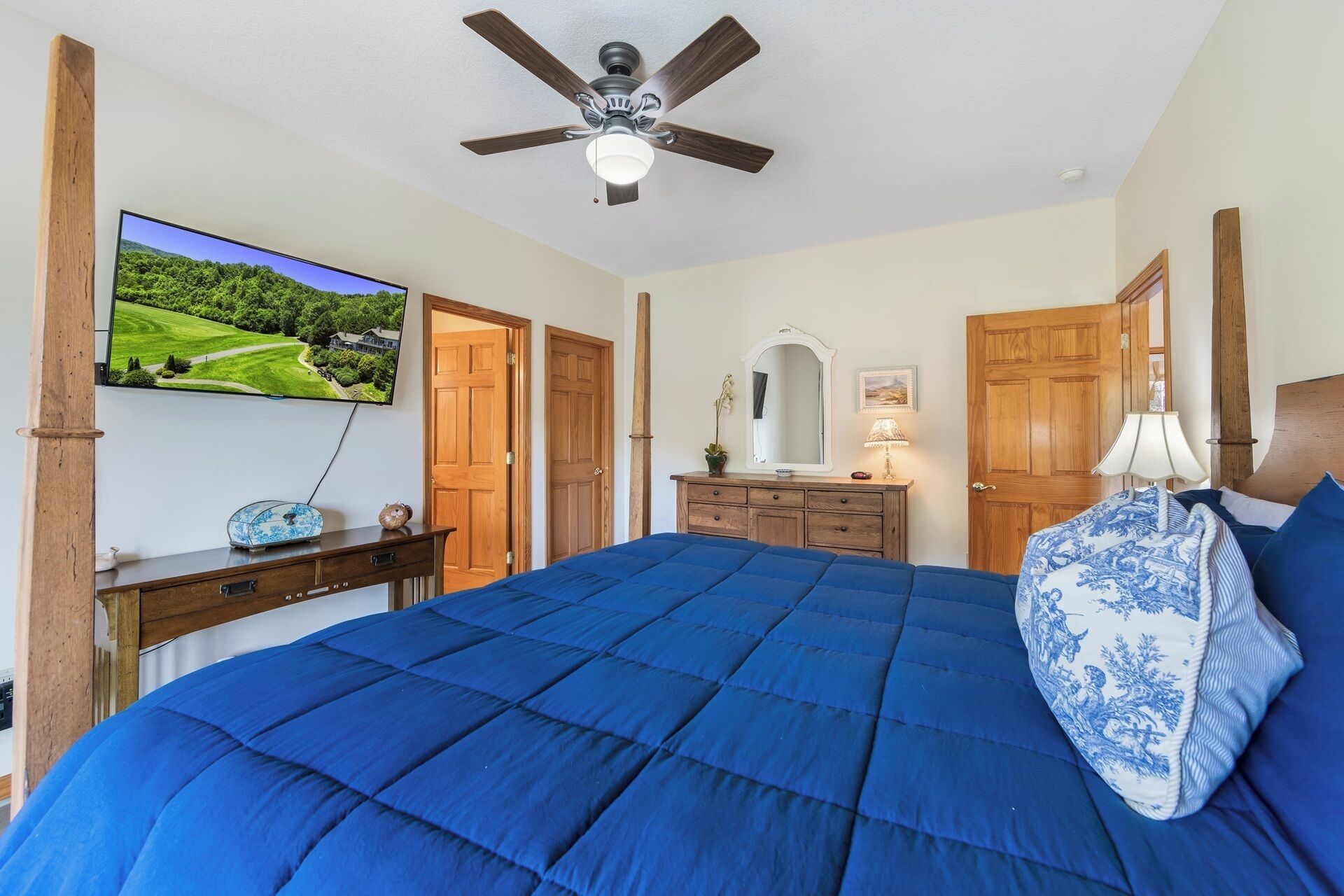 Een slaapkamer met een groot bed, een plafondventilator, een televisie aan de muur, houten deuren en een dressoir.