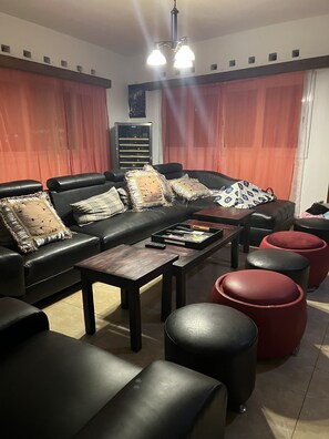 Una sala de estar con un sofá de cuero negro, una mesa de centro de madera y otomanas rojas. Hay varios cojines en el sofá, y se ve un televisor al fondo.