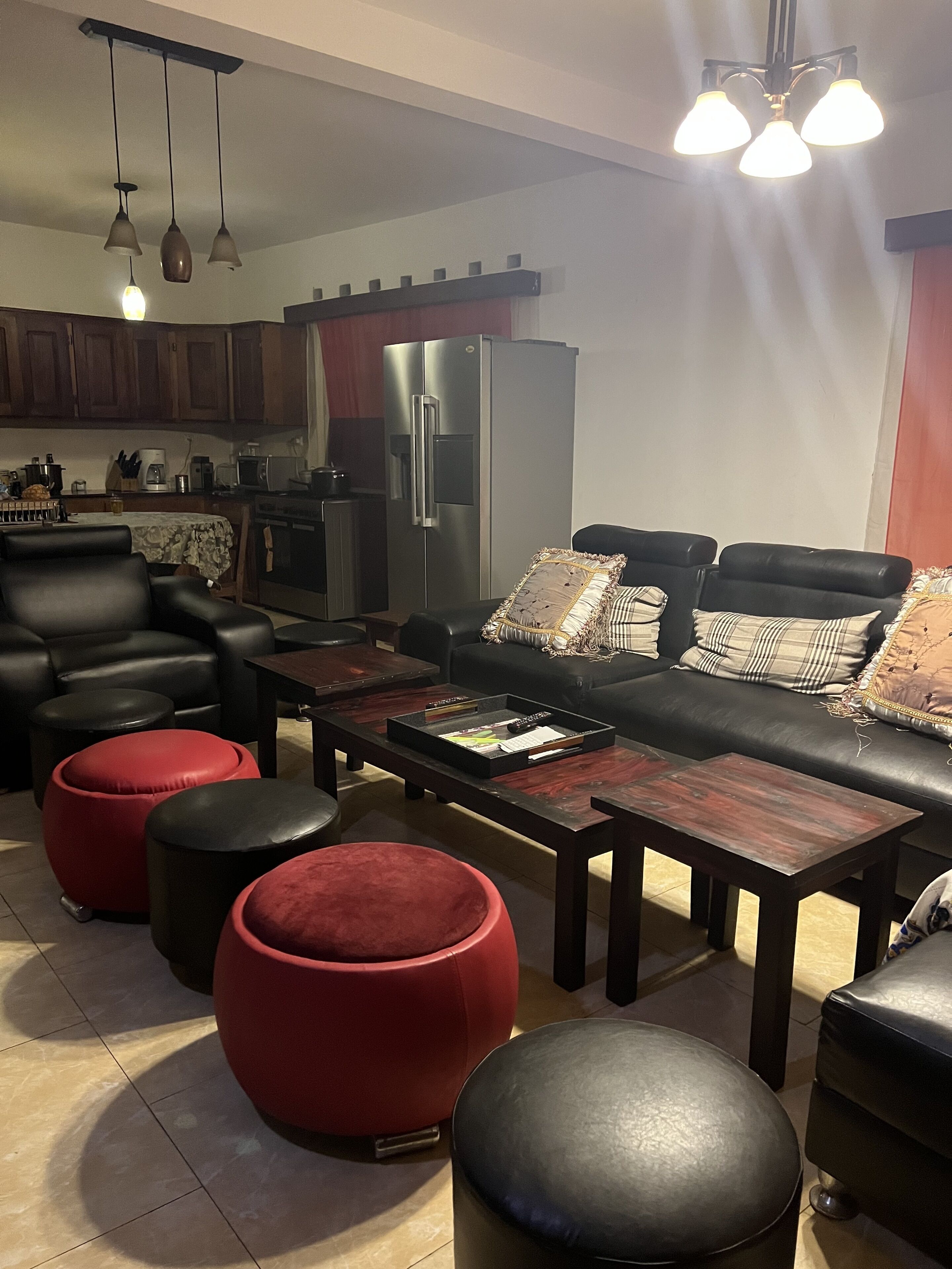 Un salon moderne avec un canapé en cuir noir, des poufs rouges et une table basse en bois. On aperçoit, au fond, une cuisine avec des meubles en bois foncé et des appareils électroménagers en acier inoxydable.