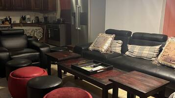 Un salon moderne avec un canapé en cuir noir, des poufs rouges et une table basse en bois. On aperçoit, au fond, une cuisine avec des meubles en bois foncé et des appareils électroménagers en acier inoxydable.